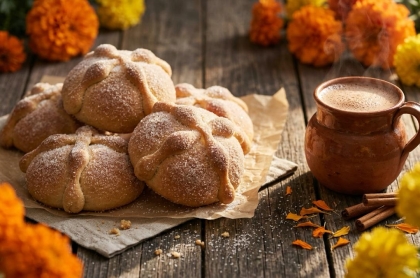 Pan de muerto espolvoreado de azúcar junto a una taza de chocolate caliente y flores de cempasúchil"