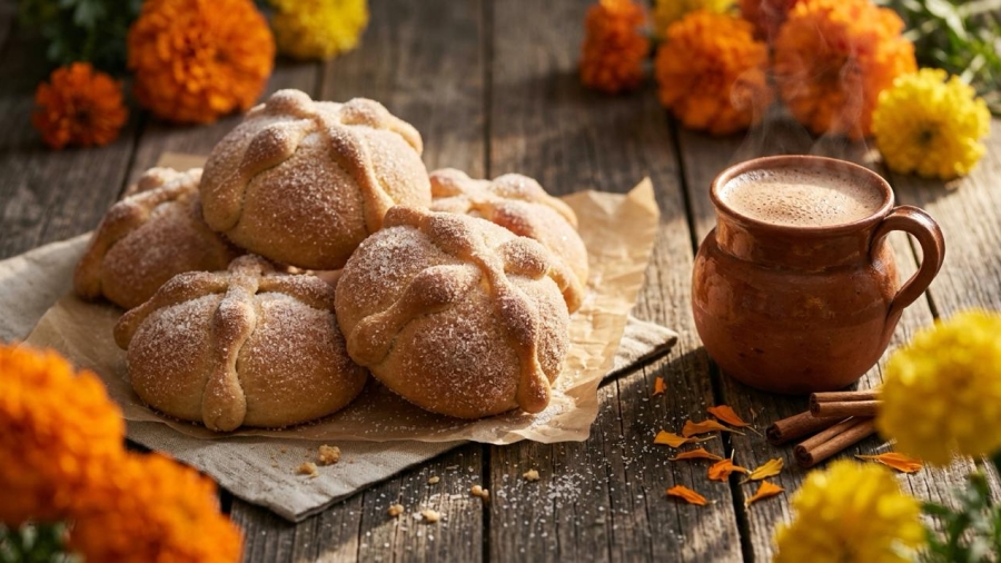 Pan de muerto espolvoreado de azúcar junto a una taza de chocolate caliente y flores de cempasúchil"